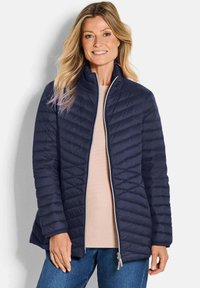 Marineblauwe gewatteerde pufferjack met hoge kraag en subtiele glans. Voorzien van een ritssluiting aan de voorkant en elastische manchetten. Geklopt over een lichtgekleurde top.