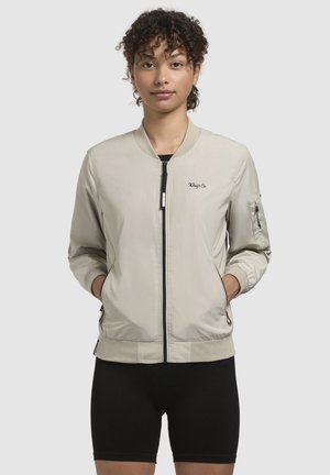 Vaaleanbeige bomber-takki, jossa on edessä vetoketju, ribbed hihansuut ja helma, brodeerattu logo rinnassa ja sivutaskut. Silkkinen kankaan pinta.