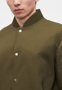 Homme portant une veste bomber vert olive avec col côtelé et boutons-pression argentés, montré du menton à la poitrine.