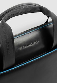 Bolso de cuero negro con cierre de cremallera, con acentos de costuras azules y un asa texturizada. Logo "Piquadro" grabado en la parte frontal.