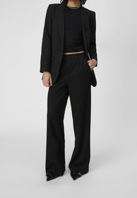 Costume noir sur mesure comprenant un blazer ajusté et un pantalon à jambes larges. Tissu lisse, design minimaliste et escarpins à bout pointu.