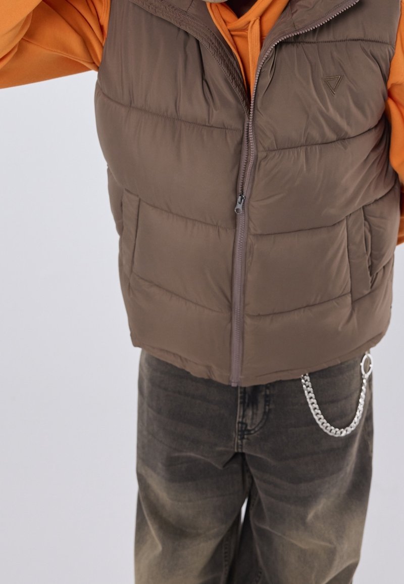 Doudoune sans manches marron avec une fermeture éclair à l'avant, un col montant et des poches latérales, portée sur un sweat à capuche orange et associée à un jean foncé déchiré.