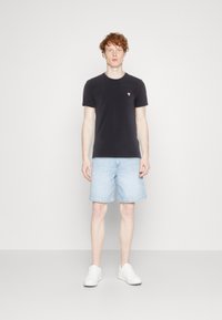 T-shirt nera di cotone con un piccolo logo triangolare rosso, abbinata a pantaloncini di jeans azzurri e sneakers bianche. Design semplice e informale.
