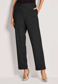 Zwarte, gestructureerde wijde broek gecombineerd met zilveren hoge hakken en een zwarte top, waarbij de linkerhand van het model in de zak zit, staand tegen een effen achtergrond.