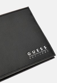 Schwarze Lederbrieftasche mit strukturiertem Oberflächen und silberner, geprägter Markenlogo "GUESS EST. 1981" in der unteren rechten Ecke.
