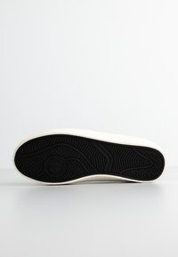 Sneaker bianco con tomaia in tessuto testurizzato e suola in gomma nera. Presenta un battistrada a motivo e un logo circolare impresso nella suola.