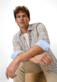 Hemd mit floralem Muster in Weiß, Blau und Gelb, mit hellblauen Manschetten, kombiniert mit hellen Hosen, sitzt auf einer strukturierten Oberfläche.