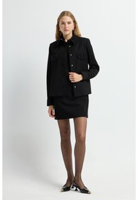Schwarze Jacke mit zwei Fronttaschen, verzierten Kragendetails, kombiniert mit einem schwarzen Kleid und transparenten Strumpfhosen. Schwarze spitze Schuhe mit goldenen Akzenten.