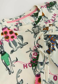 Camisa de pijama de algodón con un fondo crema claro y coloridas ilustraciones de perros vestidos con ropa navideña. Incluye acentos en verde, rosa y naranja.