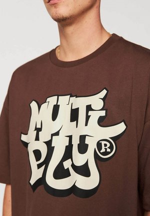 Homme portant un T-shirt marron avec un grand texte stylisé blanc et noir "MULTI PLUG" imprimé sur le devant.