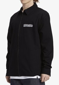 Chaqueta negra con cremallera y cuello, que presenta un logo bordado en el pecho. Está hecha de una tela texturizada, tiene mangas largas y bolsillos laterales.