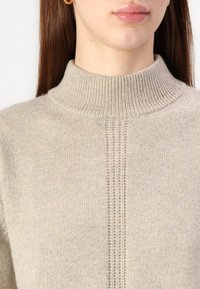 Beige stickad polotröja med ribbad krage och vertikal ribbdesign ner i mitten. Mjuk textur och varmt material.