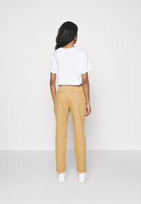 T-shirt blanc à manches courtes associé à un pantalon marron clair, avec des poches latérales et un design simple. La personne porte des baskets blanches.