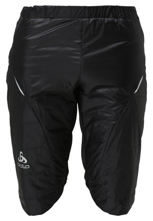 Hombre atlético con camiseta negra de manga corta, pantalones cortos negros y zapatillas de correr blancas posando contra un fondo claro sencillo.