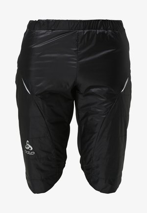 Homme athlétique portant un t-shirt noir à manches courtes, un short noir et des chaussures de course blanches posant devant un fond clair uni.