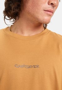 Ruskea puuvillapaita, jossa on pyöreä pääntie ja vaalenapunainen Quiksilver-logo rinnassa. Tasainen pinta ja yksinkertainen muotoilu.