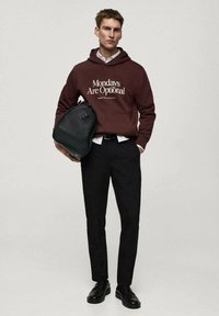 Hnědá mikina s bílým textem, černé kalhoty a černé boty. Drží texturovanou černou tašku, stojí na neutrálním pozadí.