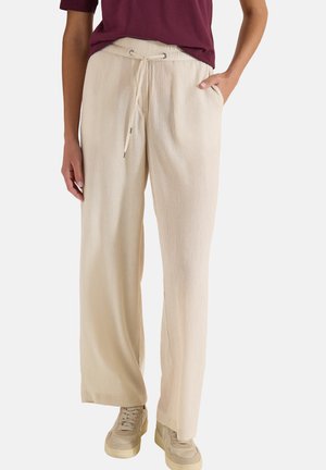 Persona con pantalones beige de pierna ancha con cintura de cordón, una mano en el bolsillo, combinados con zapatillas beige y una camiseta de color rojo oscuro.