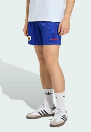 Osoba nosząca niebieskie spodenki Adidas z logo, białe skarpetki Adidas oraz białe buty sportowe z czarnymi paskami stojąca na jasnym tle.
