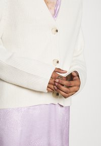 Crème kleurige ribgebreide cardigan met een V-hals, voorzien van twee grote lichtgekleurde knopen. Het model draagt een lavendelkleurige jurk eronder.
