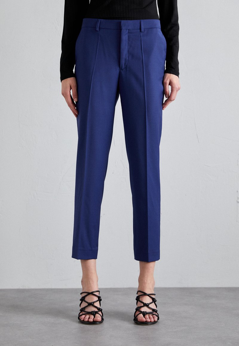 Pantaloni navy din stofă, cu croială dreaptă, având un pliu frontal și buzunare laterale, asortați cu sandale negre cu bretele.
