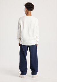 Persona con pelo rizado corto, usando un sudadera blanca, pantalones oscuros sueltos con los puños enrollados y zapatillas blancas, de pie de espaldas.