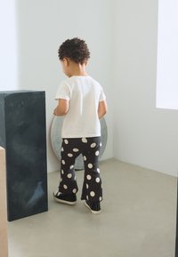 Camicia bianca a maniche corte, pantaloni neri svasati con grandi pois bianchi e scarpe da ginnastica nere. Bambino in piedi con le spalle rivolte verso lo spettatore.