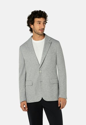 Mann trägt hellgrauen, strukturierten Blazer über weißem Hemd und schwarzen Hosen, steht vor einfachem Hintergrund und schaut zur Seite.