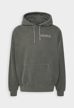 Vihreä huppari, joka on valmistettu pehmeästä kankaasta. Siinä on kengurulaukku, säädettävä nyöri ja valkoiset printtitekstit rinnassa.