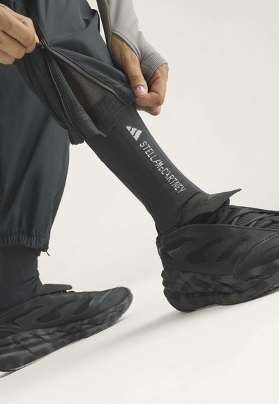 Person zieht das schwarze Hosenbein hoch, um eine schwarze Socke mit weißen Adidas- und Stella McCartney-Logos zu zeigen, trägt schwarze Sneaker.