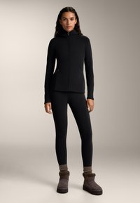 Črna zip-up jakna s kapuco, oprijete črne legice in rjave gležnjarje obložene z umetnim krznom. Gladka tekstura skozi celotno oblačilo z strukturiranim dizajnom.