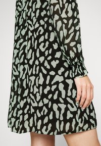 Musta mekko, jossa on mintunvihreä abstrakti kuvio, läpinäkyvä kangas, rypytetyt hihat ja väljä malli. Helman pituus on reiden keskikohtaa ylempänä.