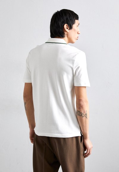 Weißes Poloshirt mit grünem Kragen, kurzen Ärmeln und subtiler Textur. Getragen mit braunen Hosen, wobei ein sichtbares Tattoo am Arm zu sehen ist.