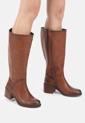 Bruine leren laarzen tot over de knie met blokhakken, gedragen op blote benen, voorzien van ritssluitingen aan de zijkant en afgeronde neuzen.