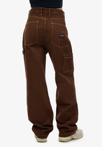 Pantalones cargo marrones hechos de un tejido resistente, con un corte relajado, múltiples bolsillos, costuras beige y una etiqueta de marca rectangular en la parte trasera.