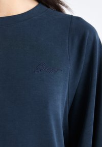 Μπλε ναυτικό πουκάμισο με μακρύ μανίκι από μαλακό υλικό, που διαθέτει ένα διακριτικό τονισμένο κεντημένο λογότυπο στο στήθος, με χαλαρή εφαρμογή και ομαλή υφή.