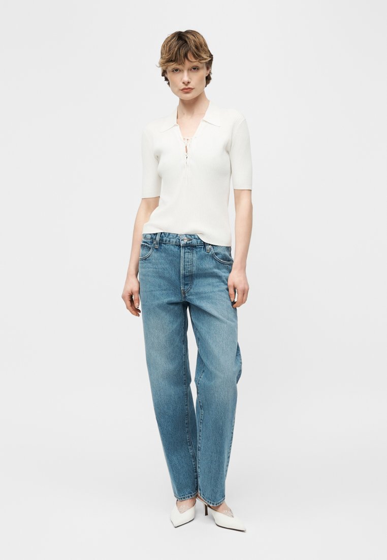 Jeune personne portant un haut en maille blanc à manches courtes, un jean bleu ample taille haute et des talons blancs pointus, debout devant un fond uni.