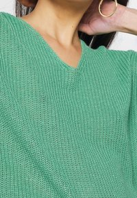 Gros plan sur une personne portant un pull en V en tricot vert et une grande boucle d'oreille en or, la main reposant près de l'oreille.