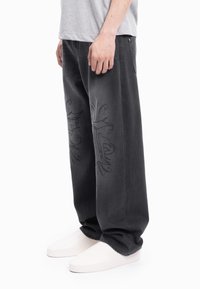 Črne široke kavbojke iz jeansa z zapletenim črnim vezenjem ob strani, kombinirane z belimi čevlji brez vezalk.