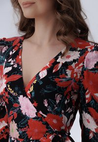 Mujer con un vestido envolvente negro con flores rojas, rosas y blancas, mostrando el escote y con cabello castaño ondulado, contra un fondo claro.