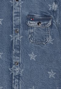 Blå denimstoff med lyse stjernemønstre, med en knappebrystlomme med metallsnappknapp og liten rød, hvit og blå merkelapp.