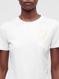 Personne portant un t-shirt blanc uni avec un petit logo circulaire Versace Jeans Couture en or clair sur le côté gauche de la poitrine.