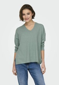 V-hals, relaxed-fit trui in zachte groene stof met lange mouwen, gecombineerd met blauwe jeans. De textuur lijkt glad en lichtgewicht te zijn.