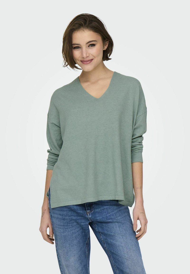 V-hals, relaxed-fit trui in zachte groene stof met lange mouwen, gecombineerd met blauwe jeans. De textuur lijkt glad en lichtgewicht te zijn.