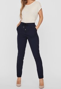 Granatowe spodnie z elastycznym paskiem ze sznurkiem, kieszeniami bocznymi i zwężanymi nogawkami, w połączeniu z jasnobeżową bluzką z krótkim rękawem i cielistymi szpilkami.