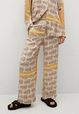 Osoba ubrana w luźne spodnie w kwiatowy wzór oraz dopasowany top z żółtymi akcentami, ubrana w czarne plecione sandały, stojąca na jednolitym tle.