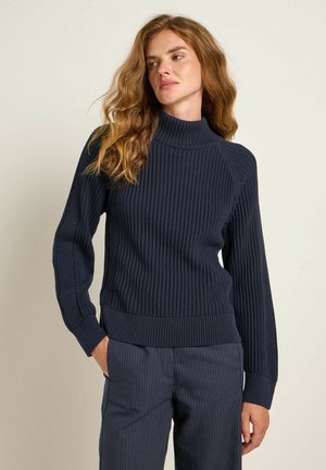 Dunkelblauer gerippter Strickpullover mit Rollkragen, langen Ärmeln und enganliegendem Saum, kombiniert mit navy- und rotgestreiften Hosen. Strukturiertes Material.