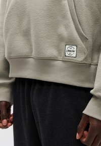 Κλείσιμο πλάνου ενός ατόμου που φοράει ανοιχτό γκρι φούτερ Nike με λογότυπο σε μπαλώμα και σκούρο παντελόνι, με τα χέρια χαλαρά στο πλάι.