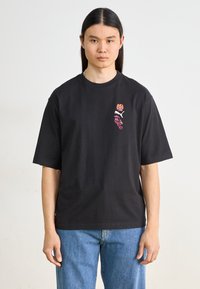 Svart bomullströja med korta ärmar, med blommig broderi i orange och rosa på vänster bröst. Rakt snitt, minimalistisk design.