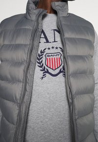 Chaleco acolchado gris con patrón de acolchado horizontal, cierre completo y cuello alzado, superpuesto a un sudadera gris que presenta un logo y bordados.